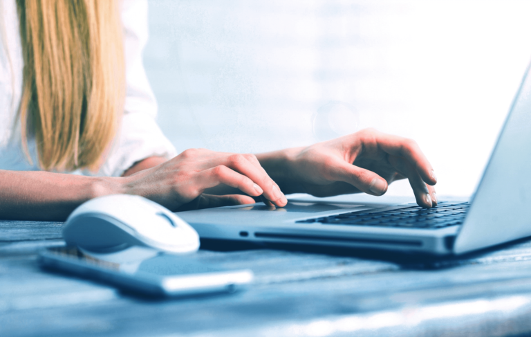 The image shows a person typing on a laptop, with a white computer mouse and a smartphone on a wooden desk nearby.