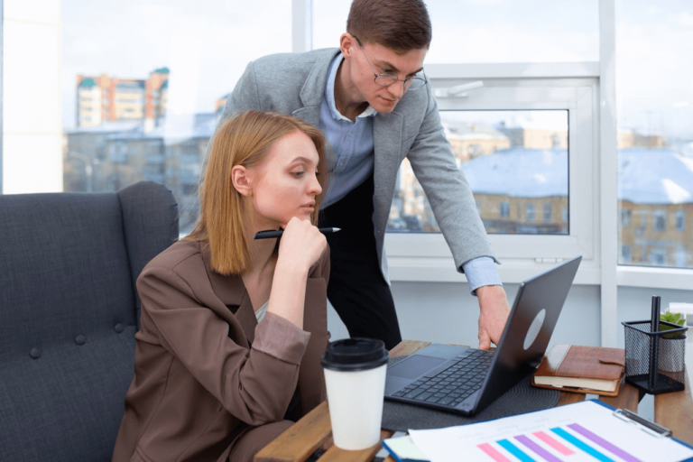 Image of a man standing beside a woman as they both look at a laptop screen, appearing to discuss something related to work.