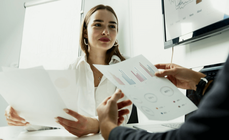 The image shows a woman in a business environment discussing topics during a meeting, while dashboards and reports with data are visible on a screen in the background.