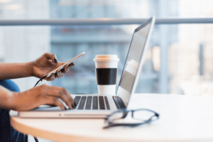 Photo of a person using a laptop and holding a phone, with a coffee and glasses next to the laptop